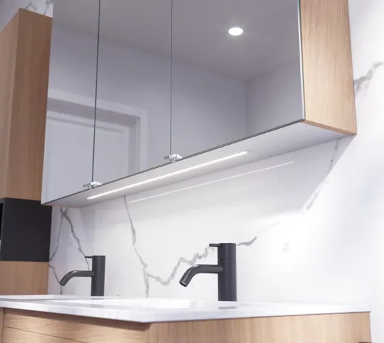 Badeværelse inspiration med vaskeskabe samt overskabe i lys eg med lameller, elegant dobbelt håndvask, smukt spejlskab med lys, sildebensgulv i lys træ og hvid marmorvæg giver et klassisk, lyst og stilrent udtryk.