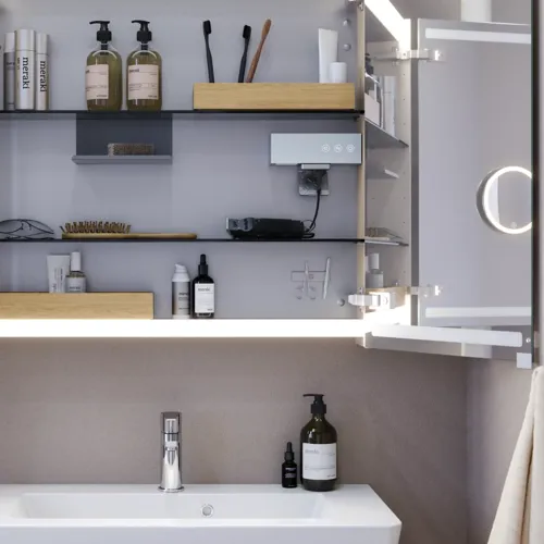 Lille badeværelse inspiration med vaskeskab og overskab over toilet i beige specialfarve, elegant spejlskab med integreret lys, de lyse farver og hvide gardin skaber en romantisk og feminin atmosfære.