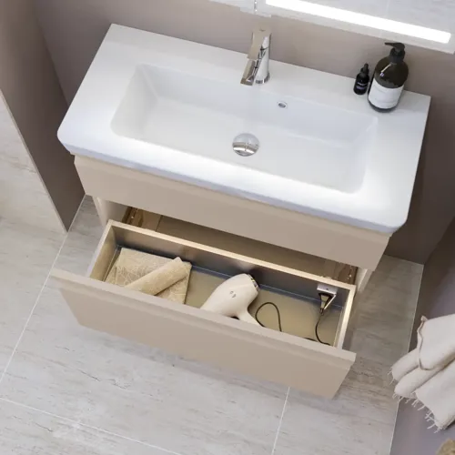Lille badeværelse inspiration med vaskeskab og overskab over toilet i beige specialfarve, elegant spejlskab med integreret lys, de lyse farver og hvide gardin skaber en romantisk og feminin atmosfære.