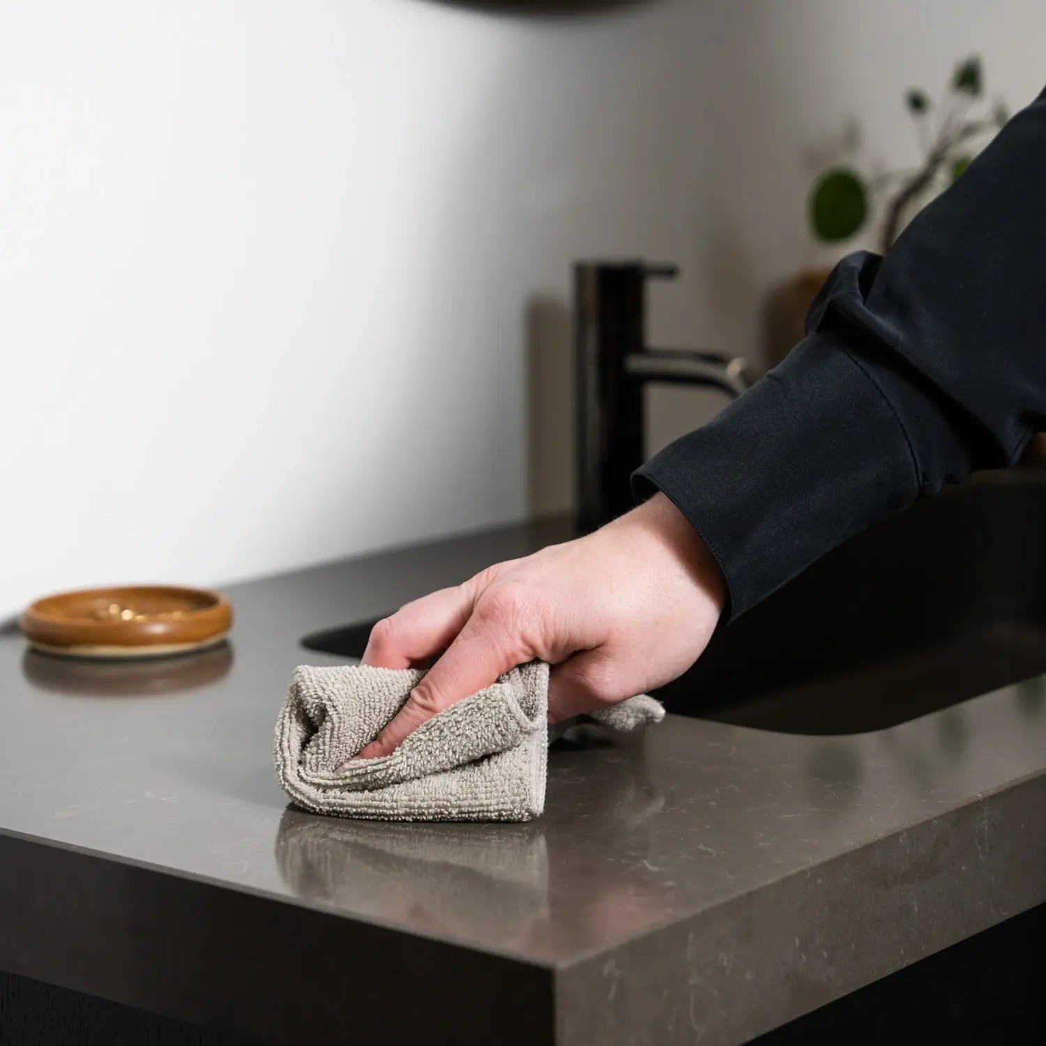 Rengøring og vedligeholdelse af badeværelsesmøbler: En person tørrer skånsomt en mørk badeværelsesbordplade af med en beige mikrofiberklud for at fjerne snavs og vandstænk.