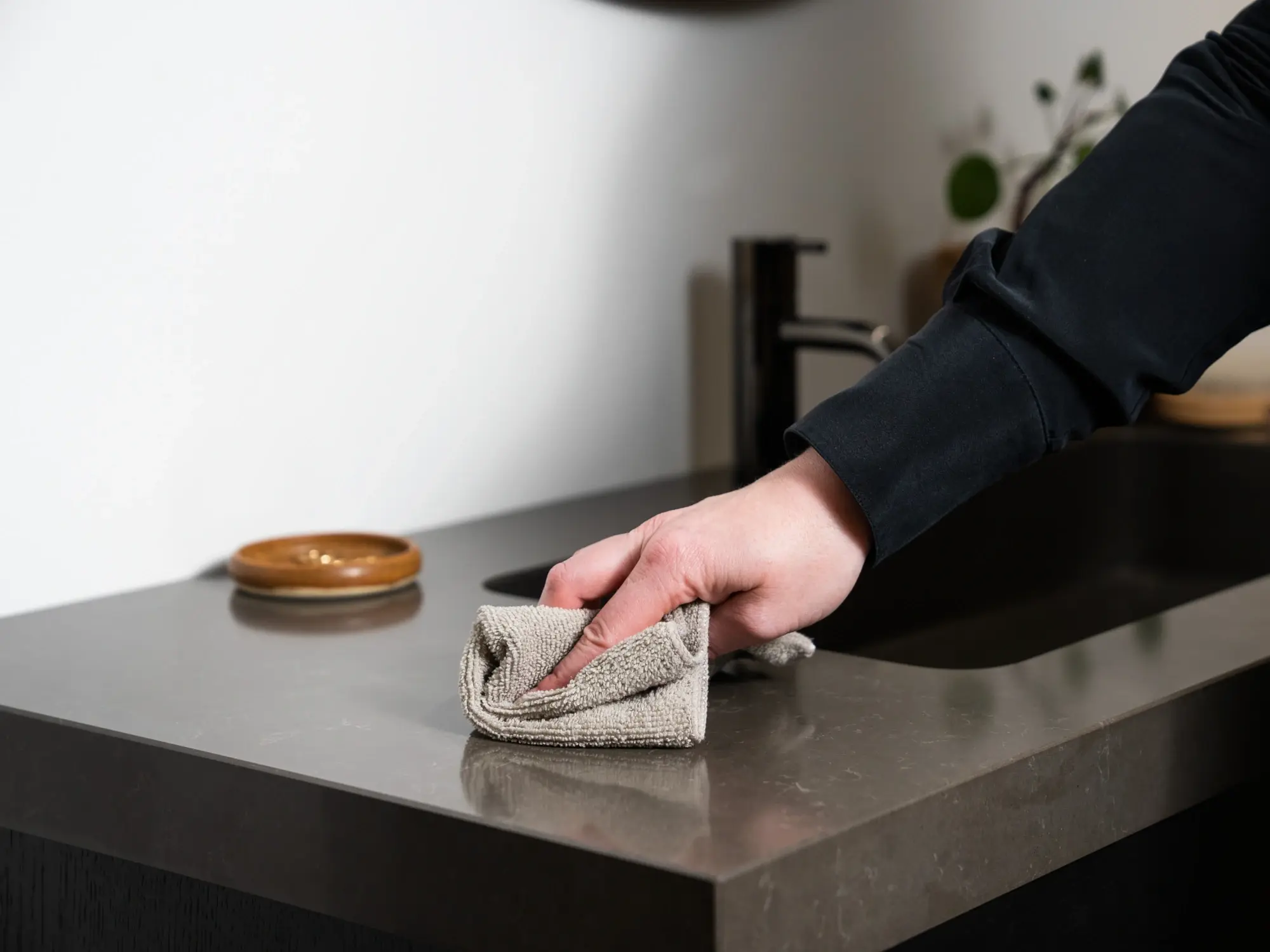 Rengøring og vedligeholdelse af badeværelsesmøbler: En person tørrer skånsomt en mørk badeværelsesbordplade af med en beige mikrofiberklud for at fjerne snavs og vandstænk.
