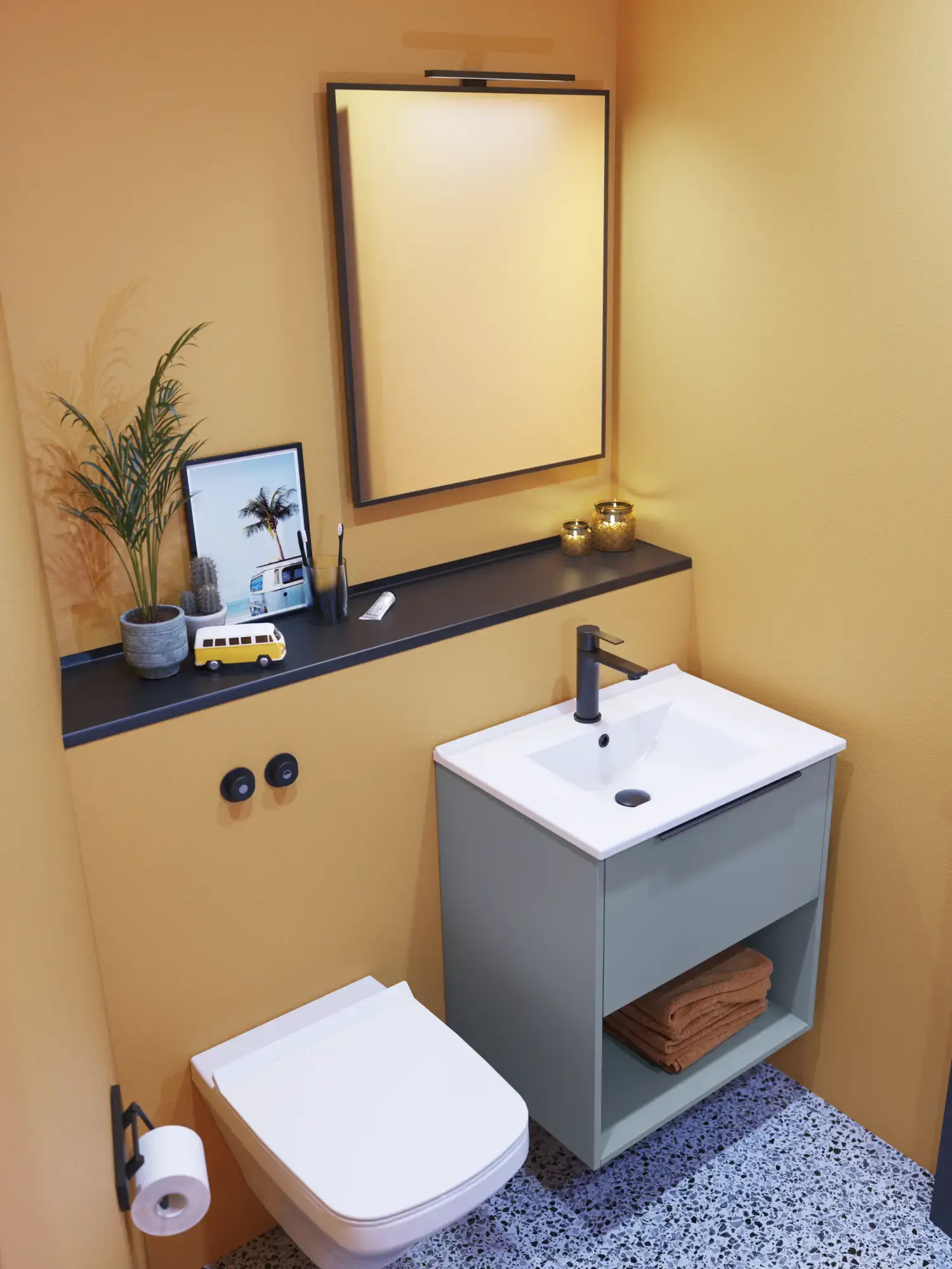 Lille badeværelse inspiration med skrå vægge og varm gule vægge, elegant vaskeskab i lys blå, tynd og stilren håndvask samt minimalistisk firkantet spejl med sort kant og praktisk lampe.